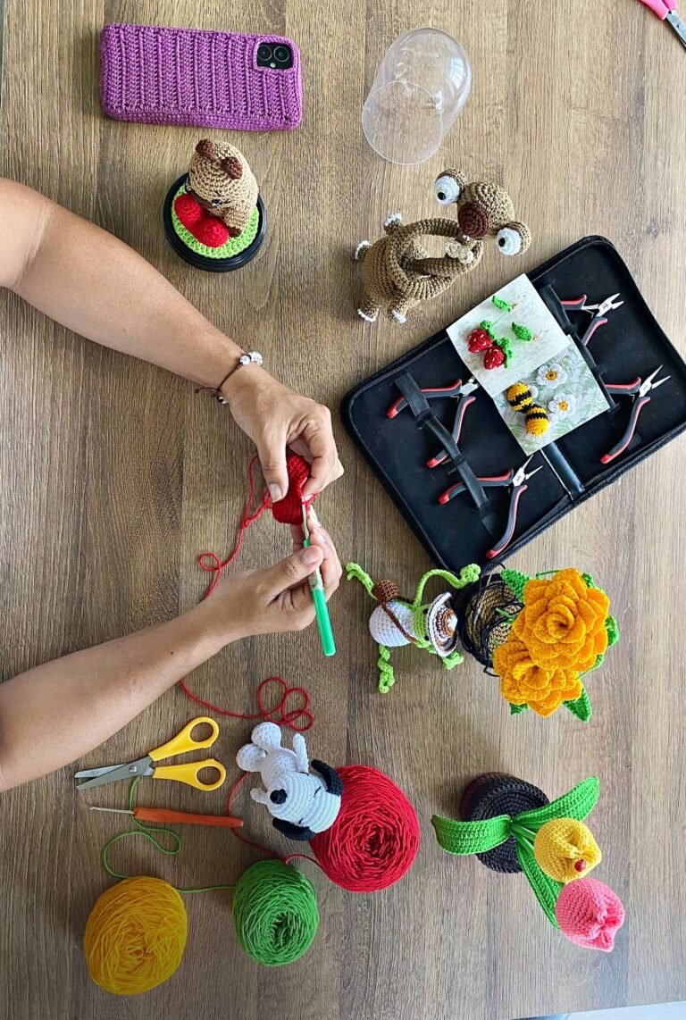Manos tejiendo a croché en la mesa del taller, entre ovillos, herramientas y piezas terminadas